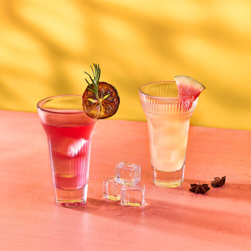 Zwei Cocktails auf rosa Untergrund mit gelbem Hintergrund, einer rot mit einer getrockneten Limettenscheibe, der andere hell mit einer Feigenscheibe, dazu Eiswürfel und Sternanis.