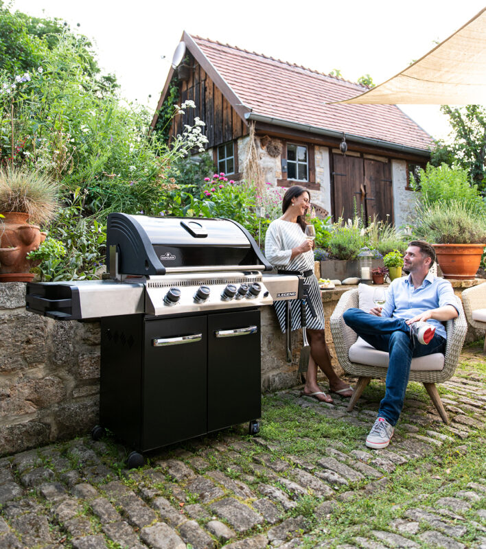 Ein Paar unterhält sich neben einem Grill in einem gemütlichen Hinterhofgarten mit einem rustikalen Holzhaus im Hintergrund.