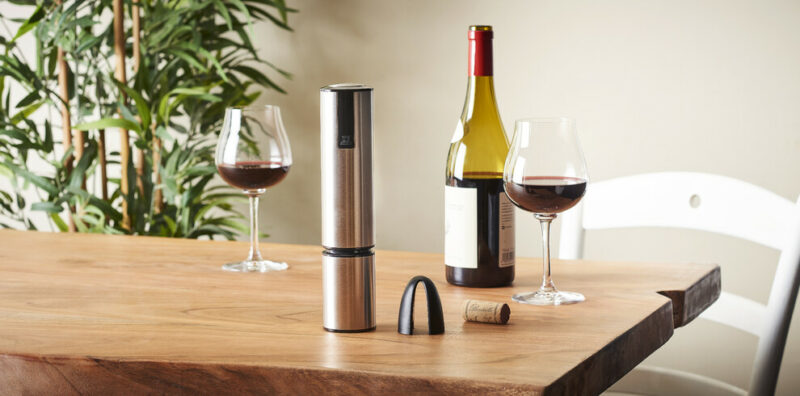 Deux verres de vin rouge, une bouteille et un ouvre bouteille électrique sur une table en bois avec une plante en arrière plan.